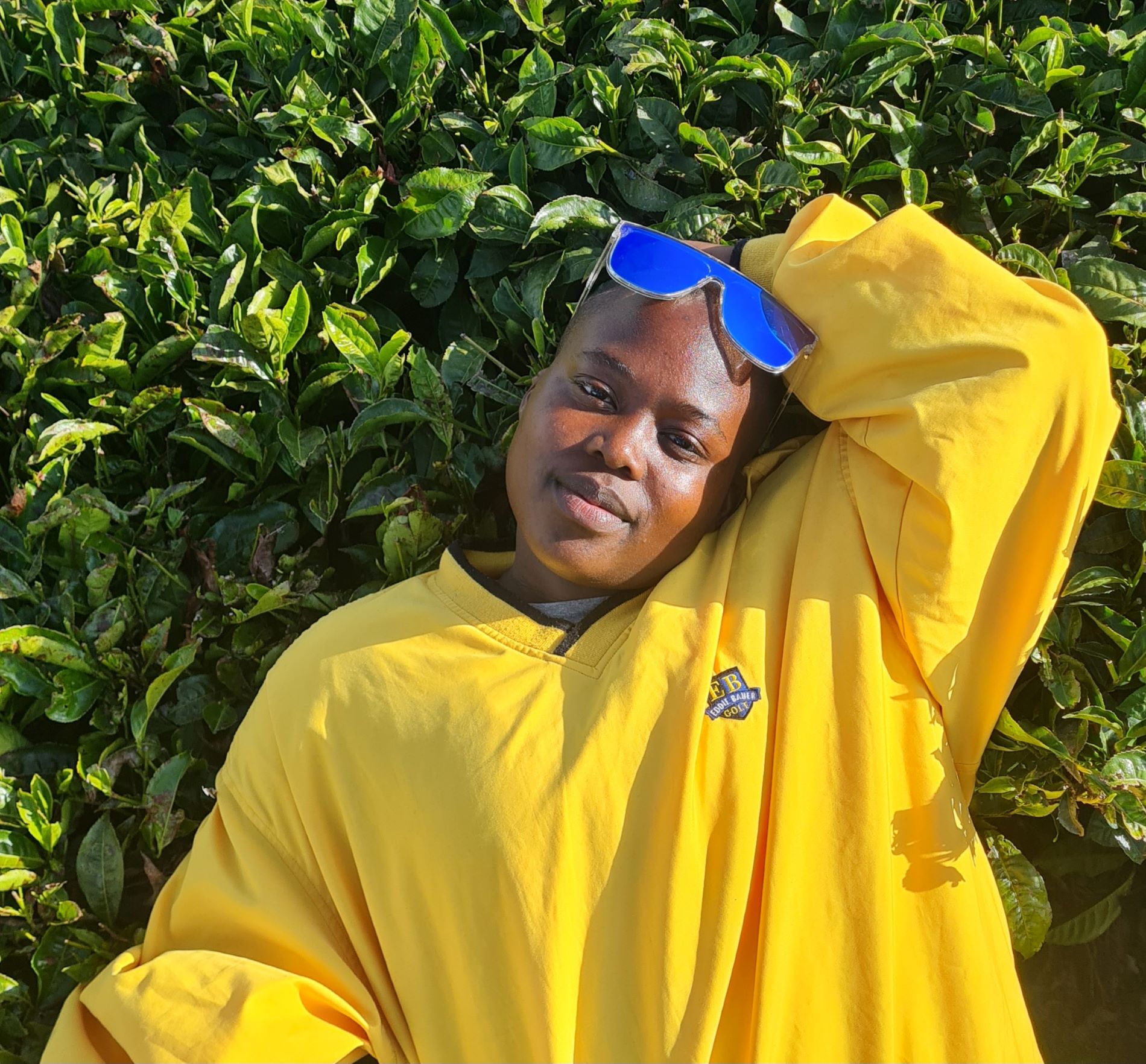 Portrait of artist Karen Wangare Leonard. She is posed in a bright yellow jacket with blue sunglasses in front of green leaves on a bush.